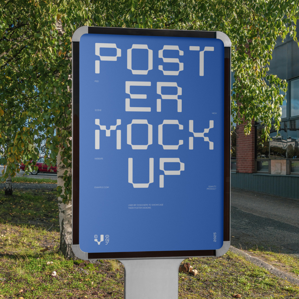 A vertical, illuminated outdoor advertising display, commonly known as a citylight poster, stands on a silver pedestal by the roadside. It features a blank white screen. In the background are green trees, a patch of grass, and a modern dark-paneled building. Free Vertical Poster PSD Mockup
