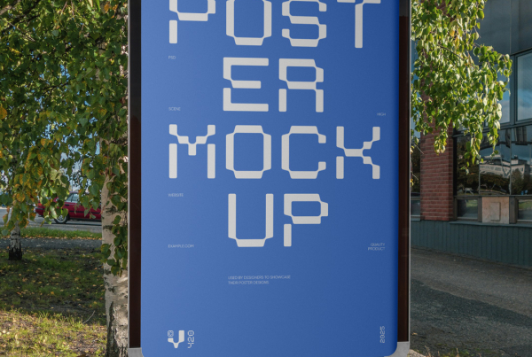 A vertical, illuminated outdoor advertising display, commonly known as a citylight poster, stands on a silver pedestal by the roadside. It features a blank white screen. In the background are green trees, a patch of grass, and a modern dark-paneled building. Free Vertical Poster PSD Mockup
