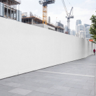 Long blank white temporary construction hoarding wall along a city sidewalk with cranes in the background.