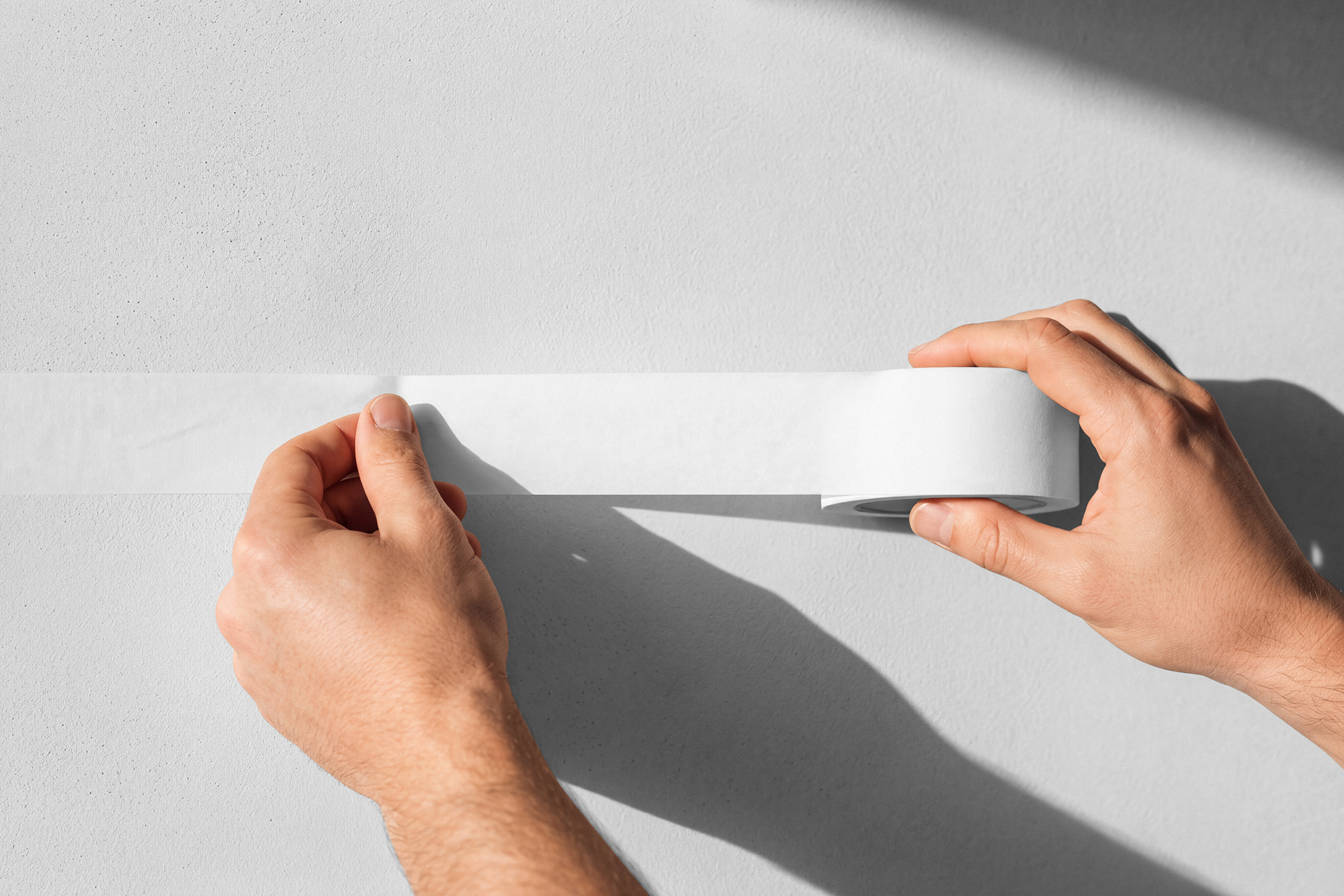 Close-up of hands applying a strip of white masking tape onto a light-colored wall.