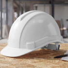 A white construction hard hat resting on an OSB table next to architectural plans and a pen.