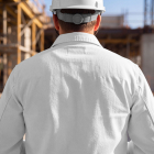 Rear view of a construction worker wearing a white hard hat and white work jacket, facing a building site.